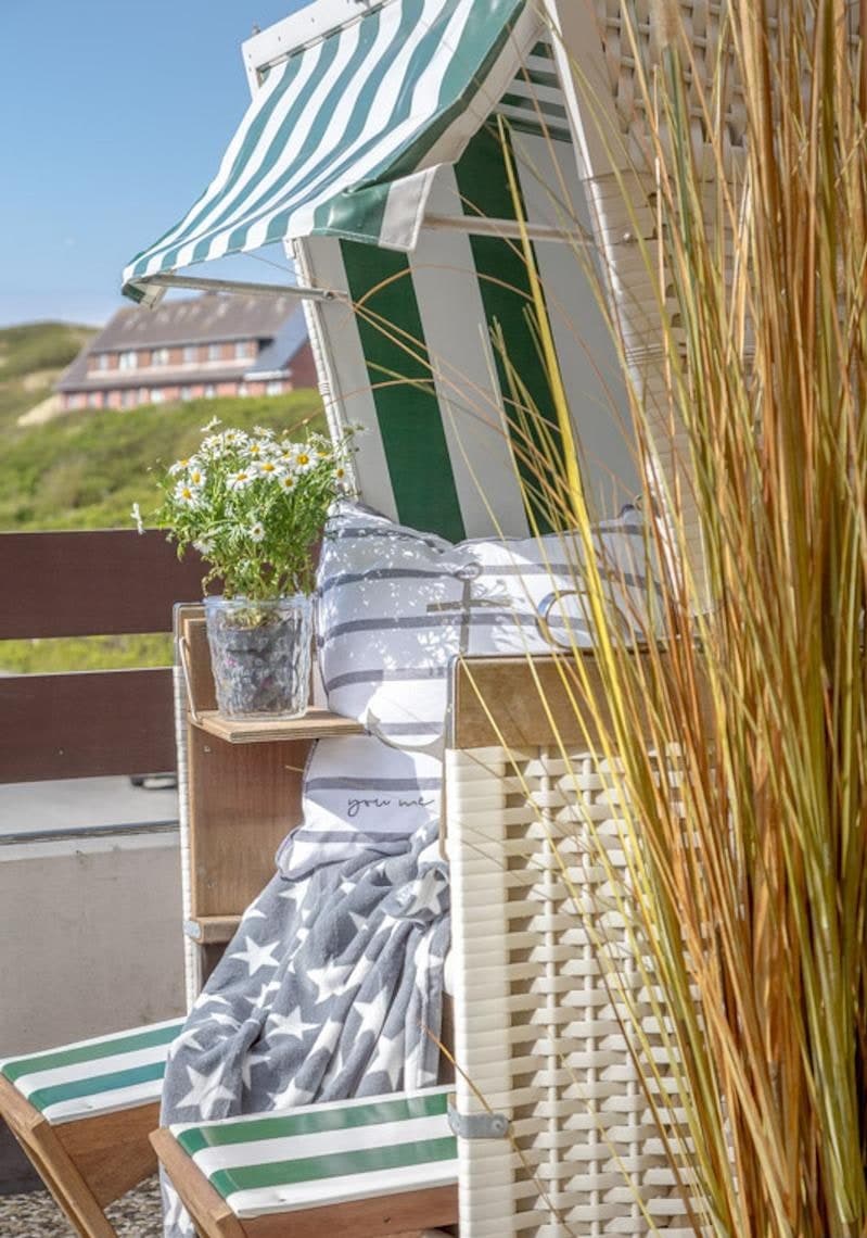 Strandkorb auf dem Südbalkon mit Kissen, Decke und Margeriten, dahinter die Dünen von Westerland und ein reetgedecktes Haus